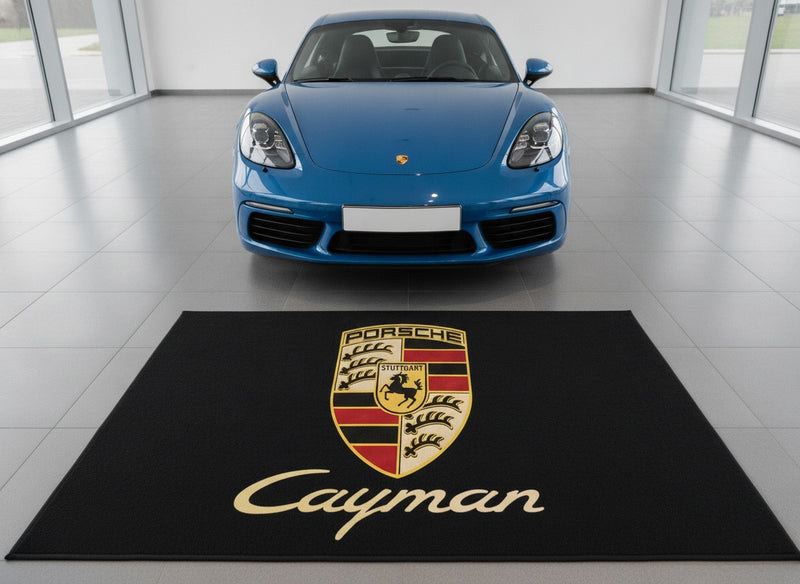 Porsche Cayman garage floor mat with crest and gold Cayman script in front of a Shark Blue 718 Cayman in a bright showroom